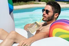 Young man wearing sunglasses drinking a refreshment on a unicorn inflatable ring in a swimming pool